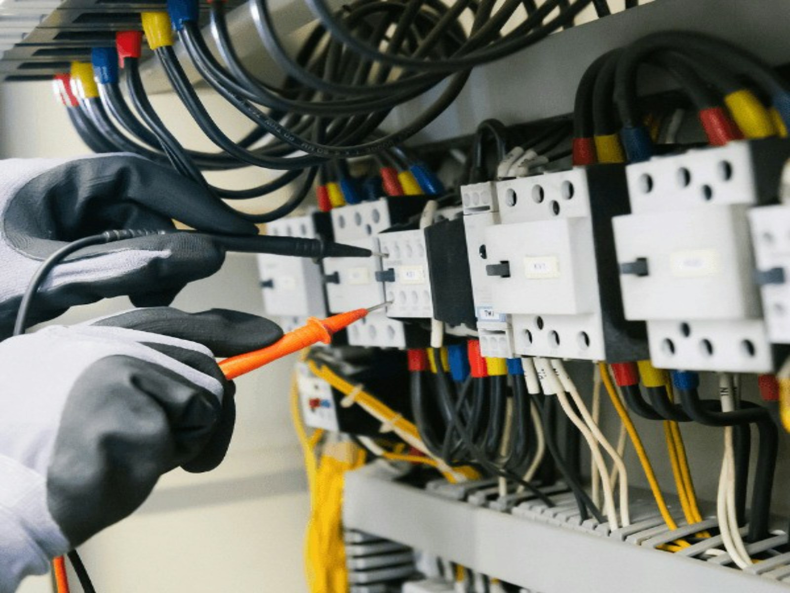 Gloved electrician probing terminals inside a load center with a multimeter, colour-coded conductors landed on breakers
