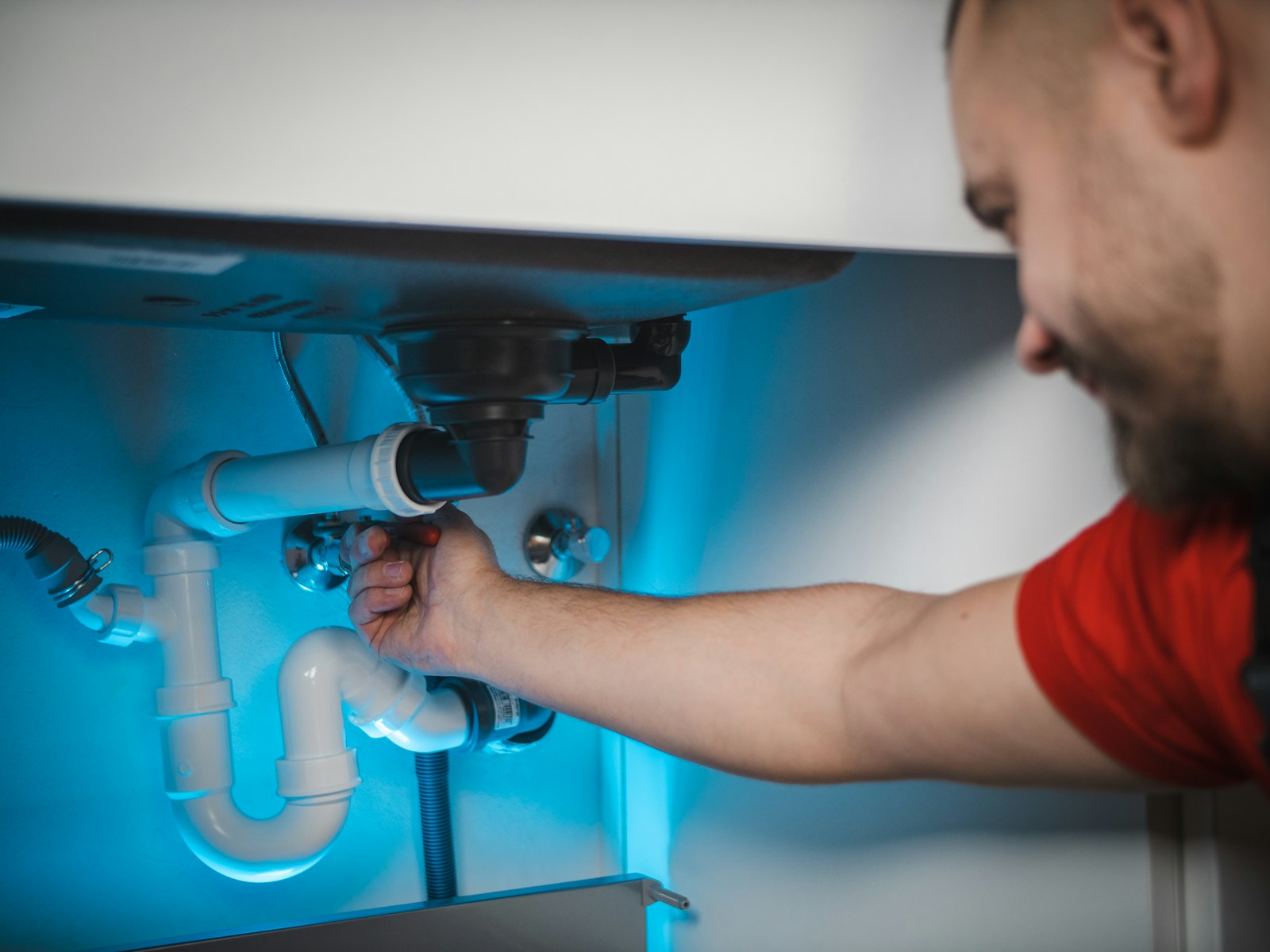 Plumber's hands tightening a supply connection on a chrome angle stop under a kitchen sink, next to a white P-trap assembly